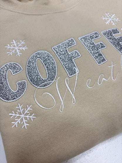 Close-up of a beige crewneck sweatshirt featuring glittery silver and white text that reads 'COFFEE' and 'We eat', decorated with white snowflake graphics.