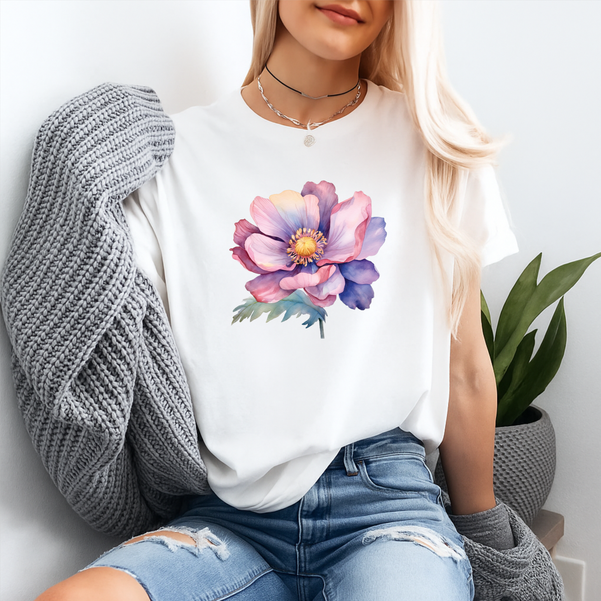 A woman wearing a white crewneck t-shirt with a colorful watercolor-style flower graphic on the front, styled with a gray knit cardigan and distressed blue jeans.