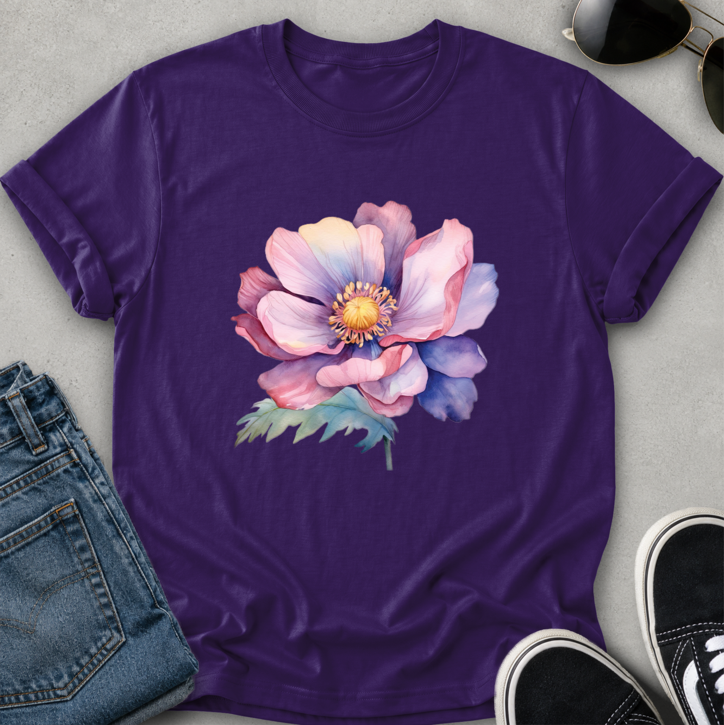 A purple short-sleeve crewneck t-shirt featuring a large watercolor-style pink and purple flower with green leaves on the front, styled in a flat lay with blue jeans, sunglasses, and black sneakers on a gray background.