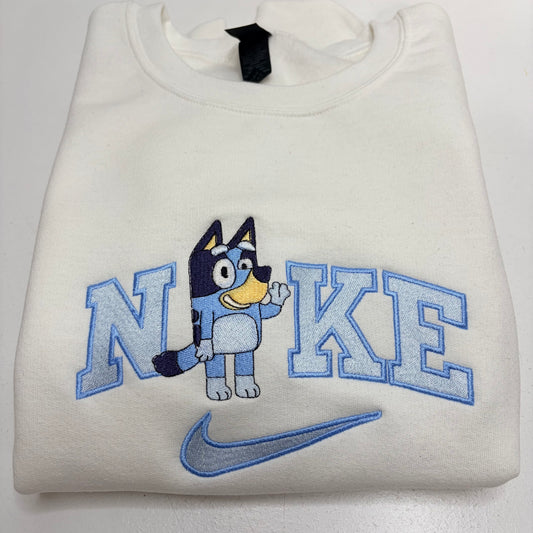 A folded off-white sweatshirt featuring a blue and navy embroidered graphic of the word 'NIKE' with a cartoon dog character in the middle and a blue swoosh below.