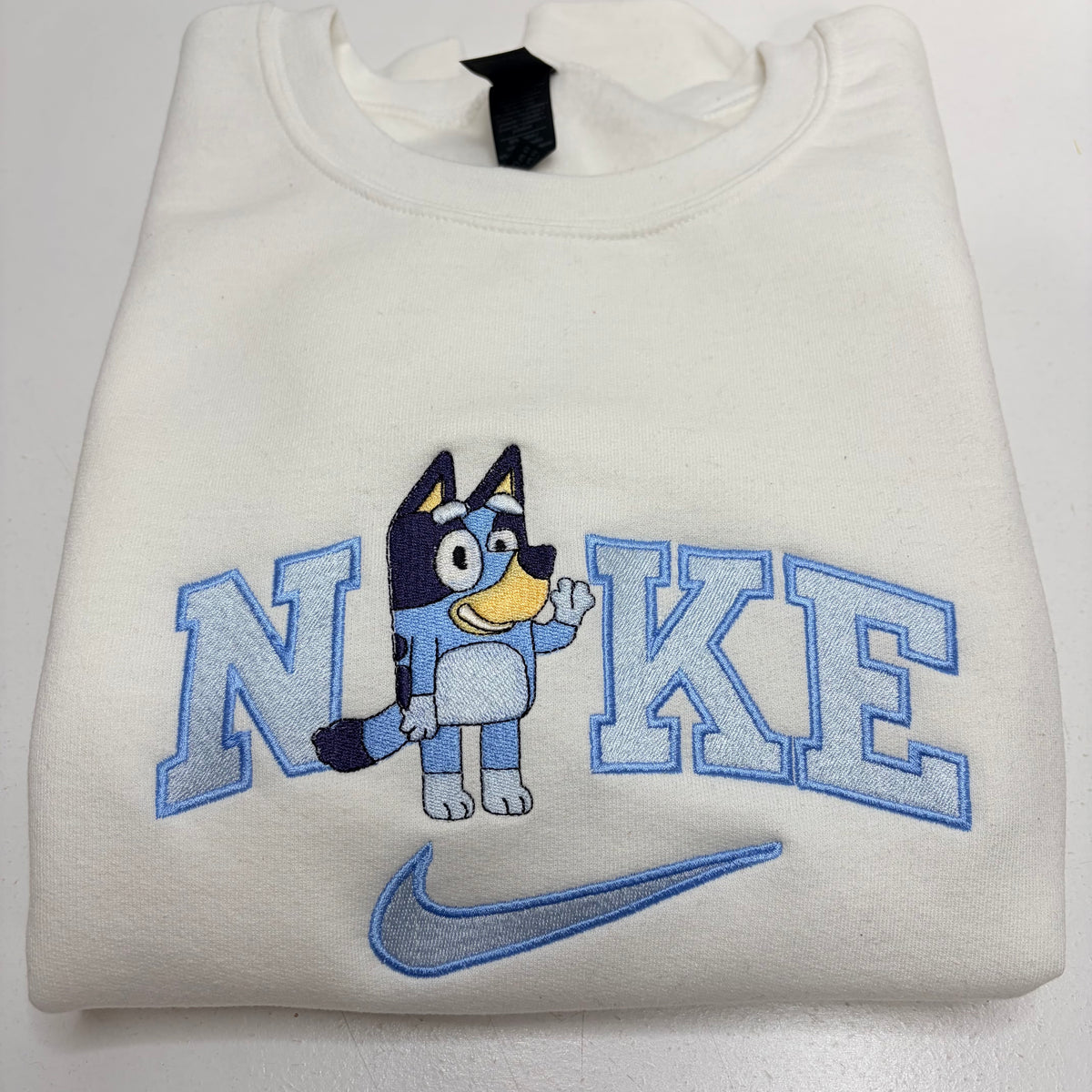 A folded off-white sweatshirt featuring a blue and navy embroidered graphic of the word 'NIKE' with a cartoon dog character in the middle and a blue swoosh below.