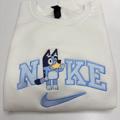 A folded off-white sweatshirt featuring a blue and navy embroidered graphic of the word 'NIKE' with a cartoon dog character in the middle and a blue swoosh below.