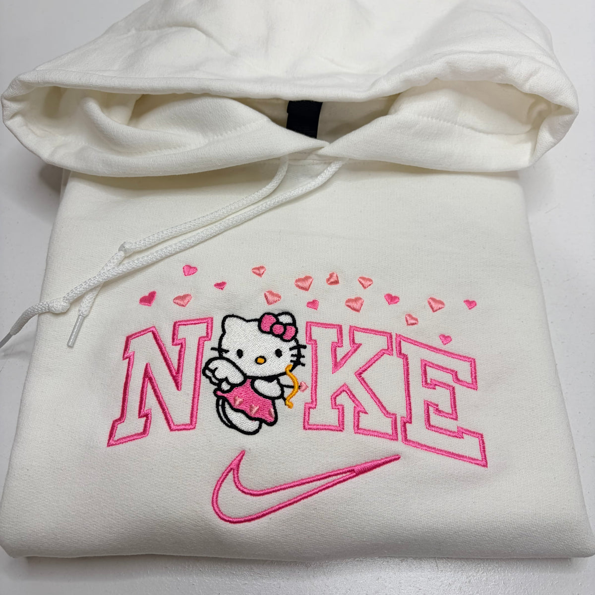 A folded white hoodie featuring a pink embroidered Nike logo, Hello Kitty character, and pink hearts on the front, with the hood and drawstrings visible.