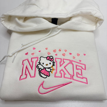 A folded white hoodie featuring a pink embroidered Nike logo, Hello Kitty character, and pink hearts on the front, with the hood and drawstrings visible.
