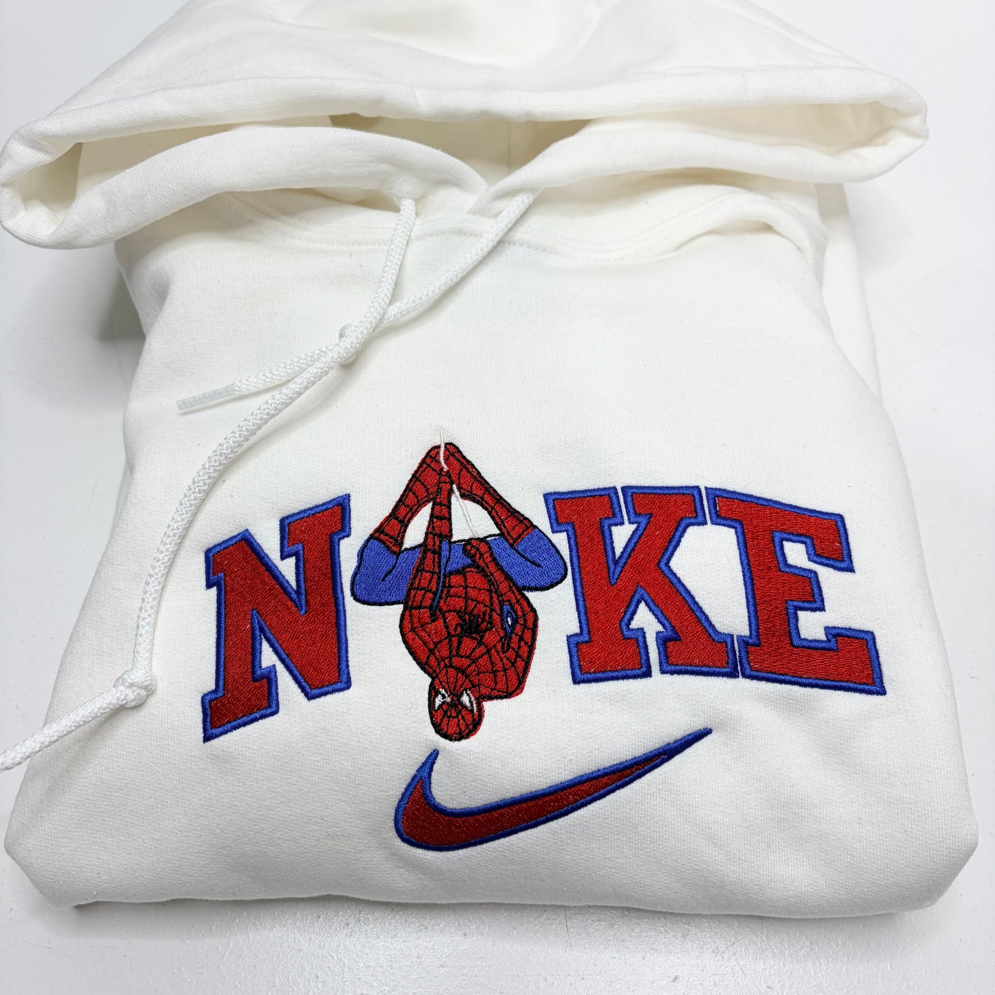 A folded white hoodie featuring a custom graphic print of the word 'NIKE' in red with blue outlines, where the letter 'I' is replaced by an upside-down Spider-Man graphic. A red and blue Nike swoosh is embroidered below the text.