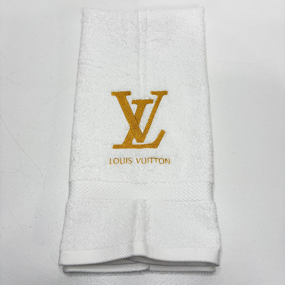 A folded white terry cloth hand towel featuring an embroidered gold Louis Vuitton LV monogram and the brand name 'LOUIS VUITTON' in gold below it, set against a plain light background.