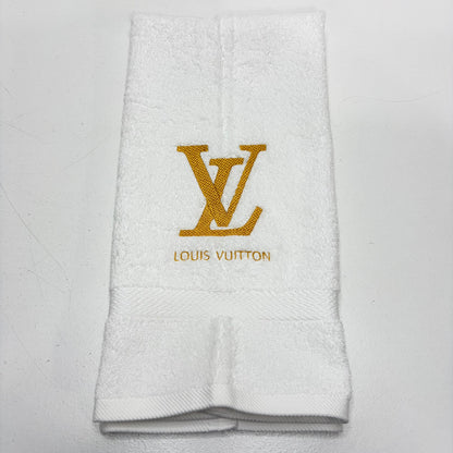 A folded white terry cloth hand towel featuring an embroidered gold Louis Vuitton LV monogram and the brand name 'LOUIS VUITTON' in gold below it, set against a plain light background.