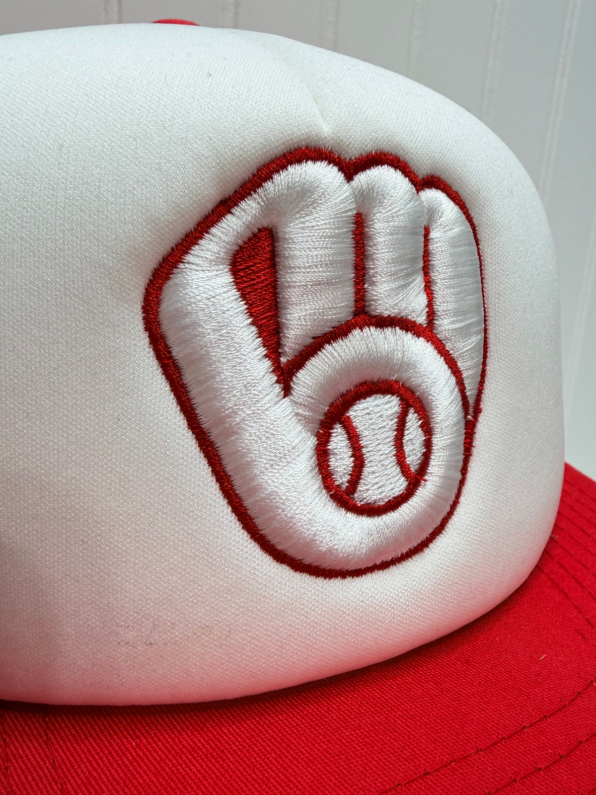 Close-up of a baseball cap's crown showing a white embroidered patch with a red outline of a baseball glove and baseball, next to red fabric.