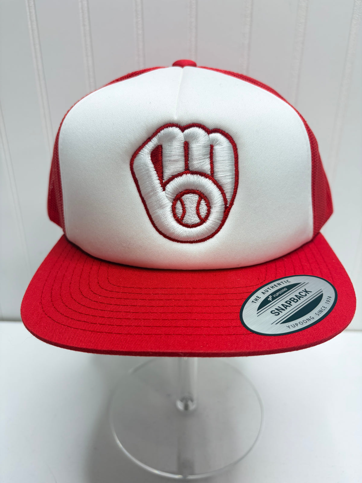 A red and white snapback trucker cap featuring an embroidered baseball glove with a baseball in the center on the front panel. The cap is displayed on a clear stand against a white paneled background.
