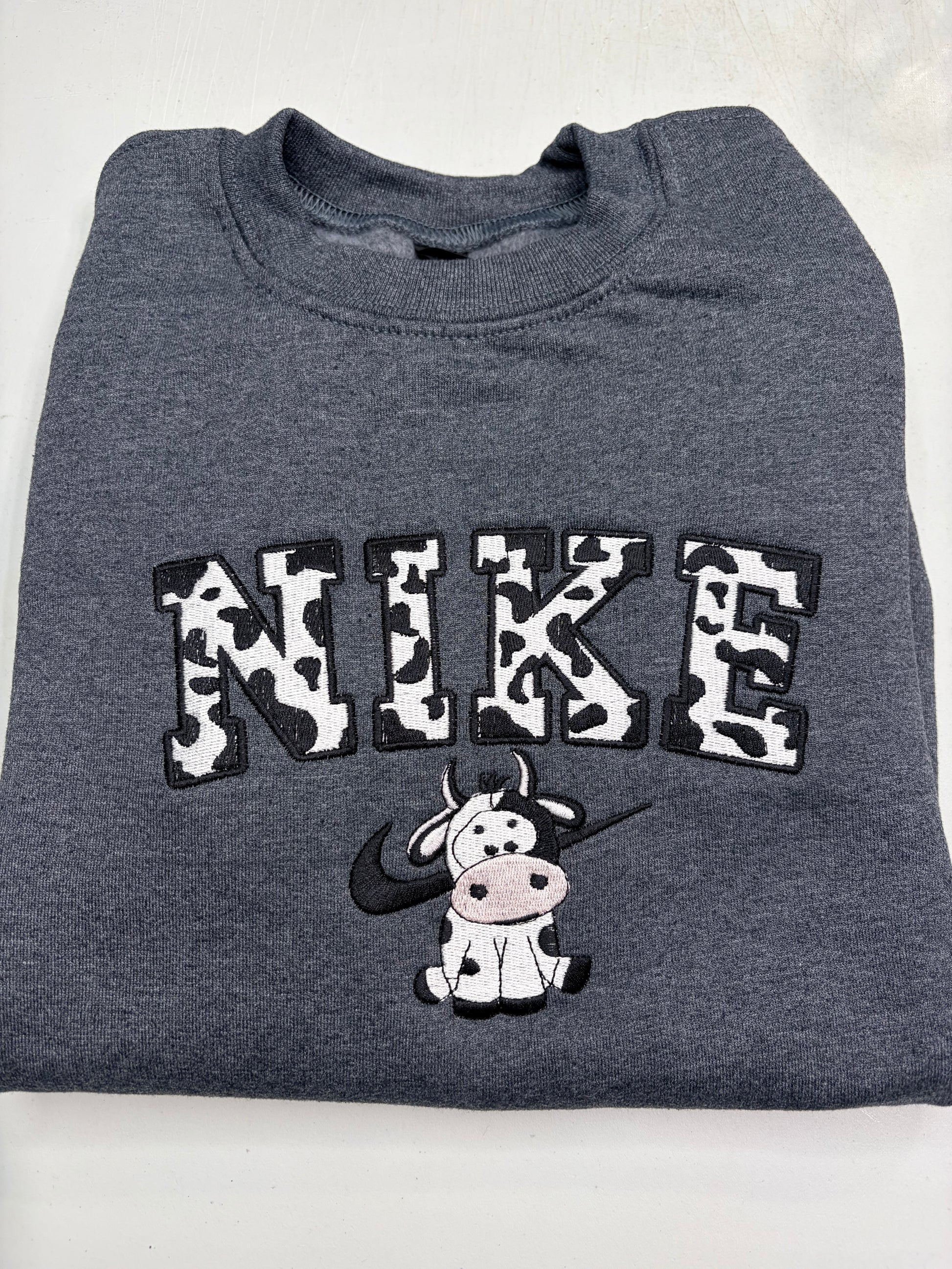 Folded gray sweatshirt with cow print 'NIKE' and cartoon cow design on a white background