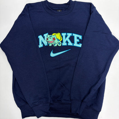 A navy blue crewneck sweatshirt featuring a 'NIKE' graphic with Bulbasaur, a light blue Nike swoosh, and a small yellow Pikachu accent, laid flat on a white background.